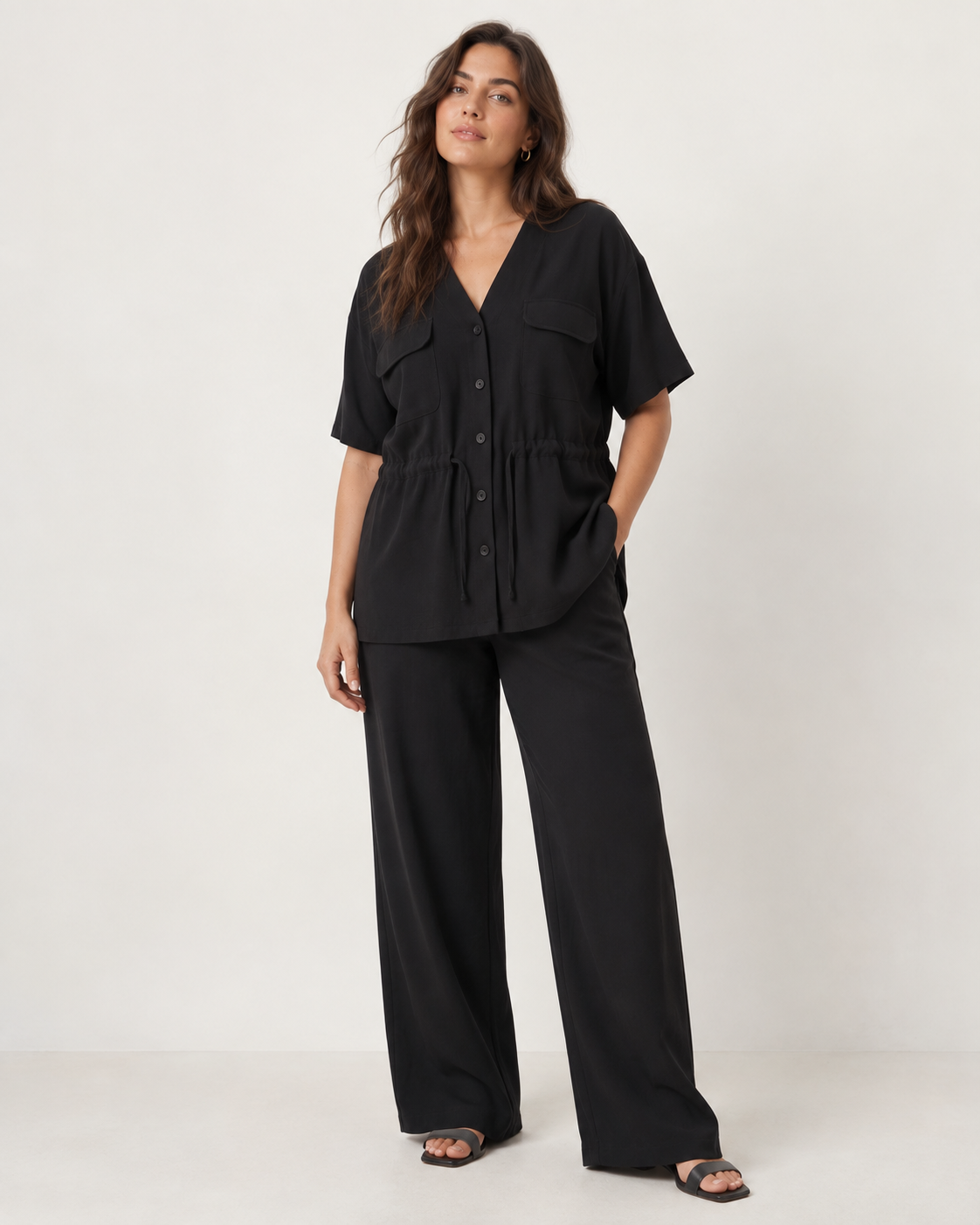 A curvy plus size curvy woman modeling a black short-sleeve button-front top with flap chest pockets and matching black wide-leg pants, photographed against a plain light studio background.