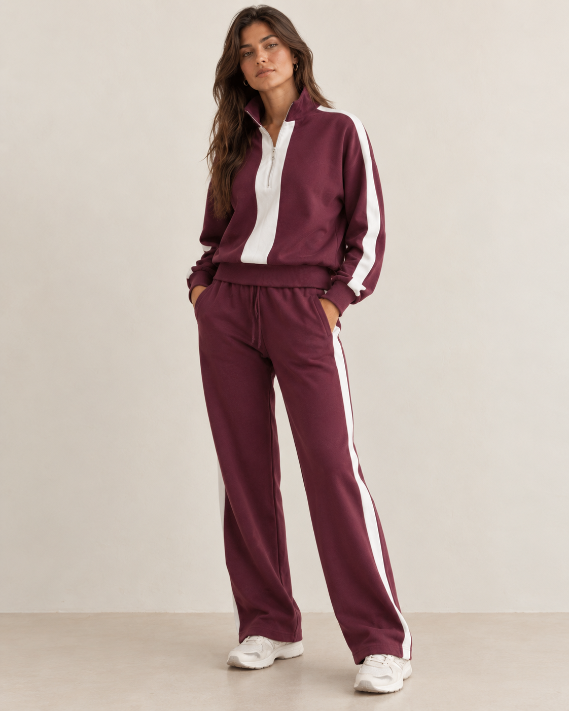 A woman modeling a matching burgundy and white tracksuit, featuring a half-zip pullover jacket and wide-leg sweatpants, set against a plain beige studio background.